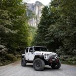 Une Jeep Wrangler robuste sur une route de gravier entourée d'une forêt luxuriante et de montagnes imposantes.